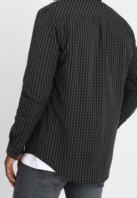 Uomo che indossa una camicia a maniche lunghe a quadri neri e grigi sovrapposta a una maglietta bianca e jeans grigi, visto da dietro.
