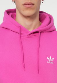 Rúžová bavlnená mikina s kapucňou so šnúrkou a bielym logom Adidas na ľavej hrudi, s hladkou textúrou a ležérnym dizajnom.
