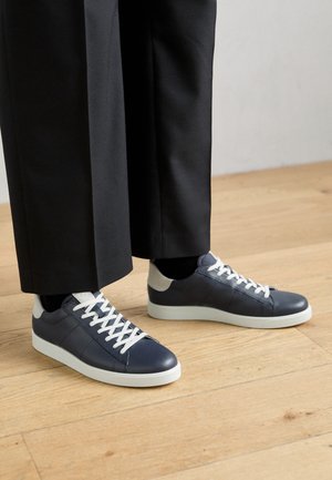 Marineblaue Leder-Sneaker mit weißen Schnürsenkeln und Gummisohlen, mit einem grauen Fersenakzent und glatter Textur, kombiniert mit schwarzen Hosen.