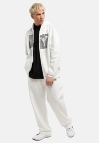 Witte hoodie met rits en zwarte grafische prints, gecombineerd met een zwart T-shirt en witte joggers. Schoenen zijn wit met zwarte accenten.