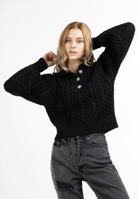 Maglione nero lavorato a maglia con motivo a treccia, dotato di colletto e tre bottoni dorati, abbinato a jeans grigi strappati a vita alta.