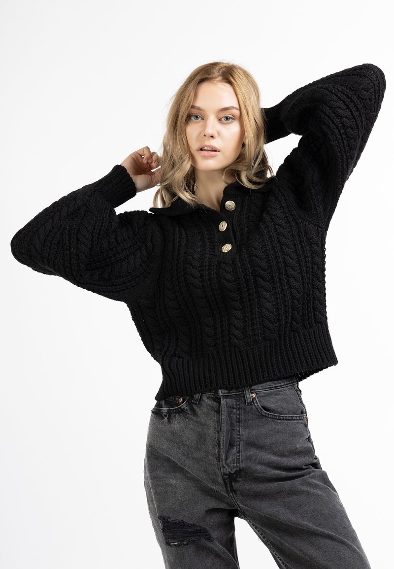 Schwarzer Strickpullover mit einem Zopfmuster, ausgestattet mit einem Kragen und drei goldenen Knöpfen, kombiniert mit hochgeschnittenen grauen Jeans im Used-Look.