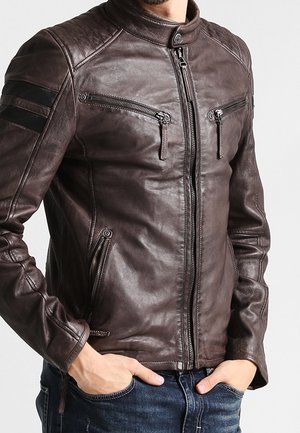 Uomo che indossa una giacca di pelle marrone scuro con tasche con cerniera sul petto e sui lati, in piedi con le mani nelle tasche dei jeans blu.