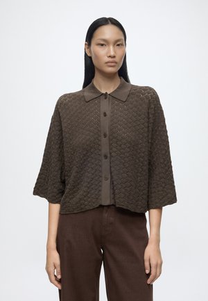 Mujer con cabello largo y negro lleva una camisa marrón oscuro con textura, abotonada y con cuello, y pantalones marrones, de pie frente a un fondo blanco liso.