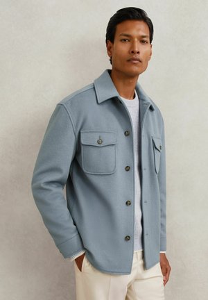 Férfi világoskék gombos dzsekiben, fehér pulóverrel és bézs nadrággal, világos egyszerű háttér előtt áll.