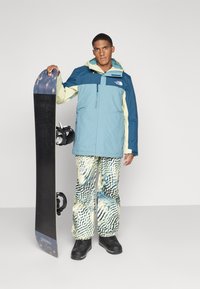 Snowboardista má na sebe vodoodolnú bundu v modrej, svetlozelenej a krémovej farbe s farebnými blokmi. Nohavice s vzorom majú vlnité línie v modrej a žltej farbe. Drží čierny snowboard.