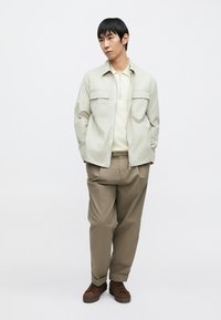 Lichtgrijs zip-up jack met twee borstzakken, gedragen over een creme blouse met kraag, gecombineerd met beige broek en bruine schoenen. Over het geheel een gladde textuur.