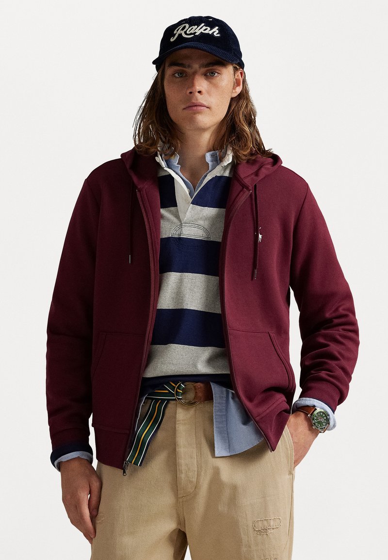 Haut à capuche zippé marron, en coton, avec poches kangourou, superposé sur un polo rayé gris et marine, associé à un pantalon beige et une ceinture marron.