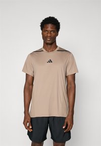 T-shirt de sport à manches courtes de couleur tan avec des accents noirs, en tissu lisse. Il présente un col rond et un logo sur la poitrine. Assorti avec un short noir.