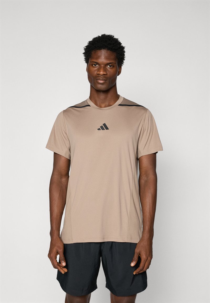 T-shirt de sport à manches courtes de couleur tan avec des accents noirs, en tissu lisse. Il présente un col rond et un logo sur la poitrine. Assorti avec un short noir.