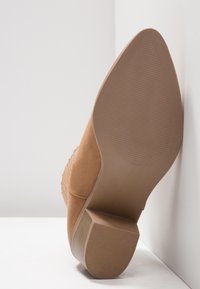 Botte pointue en daim couleur tabac avec talon bloc, vue de la semelle contre un mur et un sol blancs.