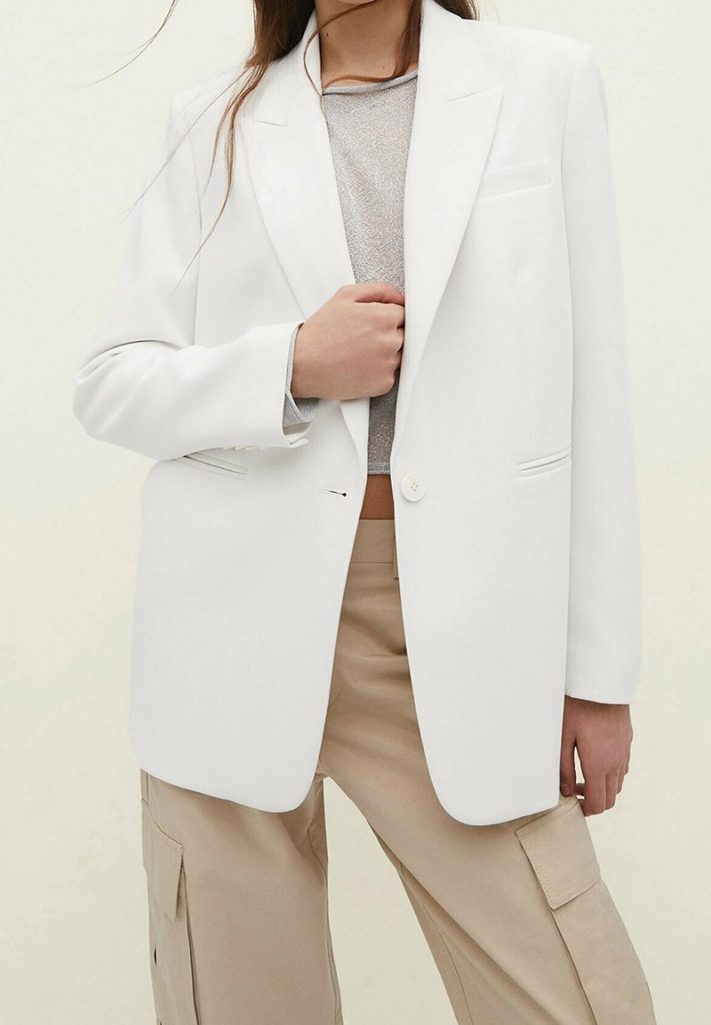 Personne portant un blazer blanc par-dessus un top court argenté et un pantalon cargo beige, la main agrippant le devant du blazer.