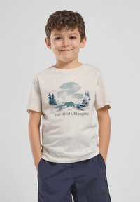 Νεαρός αγόρι με σγουρά μαλλιά, φορώντας ένα μπεζ μπλουζάκι που απεικονίζει μία αρκούδα, δέντρα, βουνά και το κείμενο "Μικροί Νομάδες, Μεγάλα Όνειρα," με τα χέρια στις τσέπες.