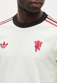 Κοντινό πλάνο ενός ατόμου που φοράει λευκό πουκάμισο με μαύρο γιακά, κόκκινες και μαύρες ρίγες στους ώμους και κόκκινα κεντημένα λογότυπα στο στήθος.