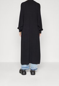 Cardigan lungo nero a maglia con maniche a palloncino, caratterizzato da una trama a costine. Indossato sopra jeans blu con orlo sfrangiato e scarpe nere chunky.