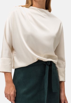 Frau trägt eine cremefarbene Satinkleidbluse mit dreiviertellangen Ärmeln, die in einen hoch taillierten dunkelgrünen Rock mit Stoffgürtel gesteckt ist.