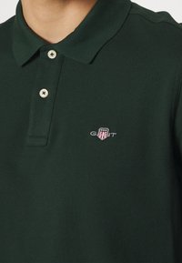 Dark green polo shirt with a textured fabric, featuring a collar, three buttons, and a small embroidered GANT logo with red and white accents.