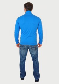 Blauwe herenpullover met een hoge kraag, gemaakt van een gladde stof. Gecombineerd met donkerdenimjeans met achterzakken en een rechte pijp.