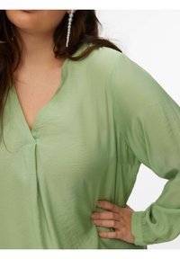 Blouse vert clair à col en V, texture douce et manches longues, avec un détail subtil de pli près de l'encolure.