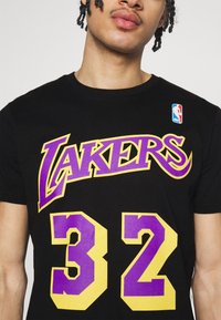 Camiseta de algodón negra con "LAKERS" en texto púrpura y amarillo y el número "32" en los mismos colores, con un parche del logo de la NBA en el hombro.