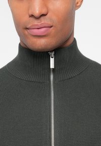 Pull en tricot côtelé vert foncé avec un col haut et une fermeture éclair à l'avant, doté d'un tirette en métal argenté et d'une surface texturée.