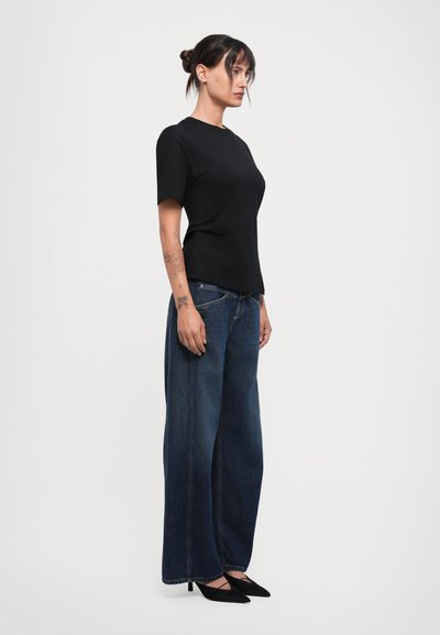 Black short-sleeve t-shirt paired with high-waisted wide-leg dark denim jeans and black pointed-toe heels. Model shown in profile view.