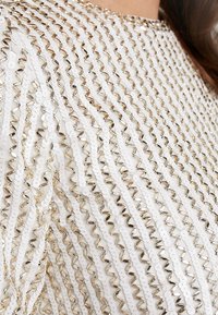 Vit tyg med paljetter och metalliska detaljer skapar ett texturerat, vågigt mönster. Ytan har växlande släta och glänsande detaljer.