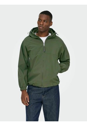 Homme portant une veste à capuche verte zippée sur un t-shirt blanc et un jean bleu, debout avec une main dans la poche sur un fond uni.