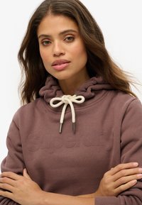Sudadera burdeos con un logo texturizado, cordones ajustables blancos y un corte holgado. Cuenta con una capucha fruncida y una tela suave y lisa.