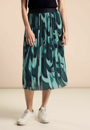 Woman wearing a pleated midi skirt with blue and black abstract patterns, a black top, and white sneakers standing on a light floor.