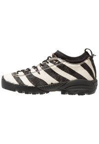 Zapatilla deportiva a rayas blancas y negras con parte superior texturizada similar a piel, suela de goma negra y diseño con cordones. Incluye una lengüeta de tirador en el talón.
