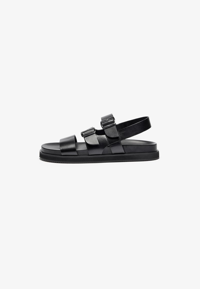 Schwarze Ledersandalen mit zwei Schnallenriemen und einem Riemen hinten, ausgestattet mit einer flachen Sohle und einer strukturierten Gummisohle für besseren Grip.