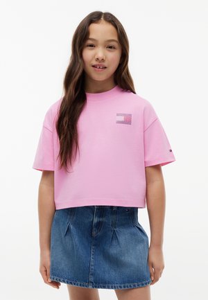 Fille aux longs cheveux bruns portant un t-shirt court rose et une jupe en jean bleue, debout devant un fond blanc uni.