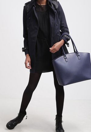 Marineblauwe trenchcoat over een zwarte jurk, gecombineerd met zwarte panty's en veterschoenen; met een gestructureerde marineblauwe tote bag.