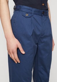Marineblauwe broek van gladde stof, met een zijriem met een gouden gesp, een knoopsluiting en een klein voorvak met een oogje.