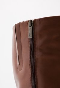 Botte en cuir marron à hauteur de genou avec fermeture éclair latérale ; présente un design épuré et des coutures discrètes. Les accessoires ont une finition métallique.