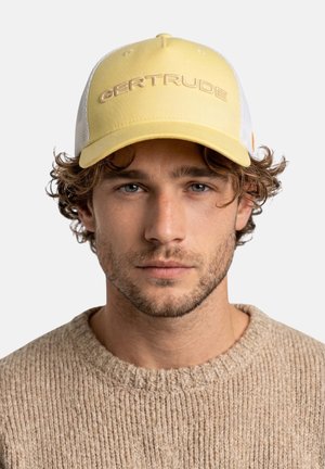 Jeune homme aux cheveux bouclés portant une casquette jaune et blanche avec l’inscription « GERTRUDE » et un pull en tricot beige, faisant face à l’avant avec une expression neutre.