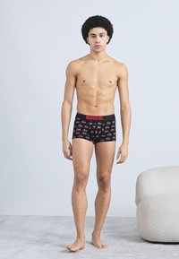 Fekete pamut boxernadrág piros derékpánttal, színes kis grafikákkal díszítve. A modell mezítláb áll világosszürke felületen.