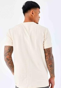Camiseta corta de manga corta beige con una costura central en la parte posterior. Presenta un corte relajado y está hecha de un suave tejido de algodón. μονιμοποιησε το χαος.