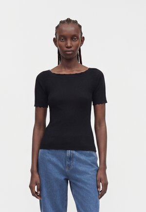 Femme aux cheveux tressés portant un haut noir côtelé ajusté à manches courtes et un jean bleu taille haute, debout devant un fond uni.