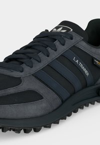 Μαύρο και σκούρο γκρι αθλητικό παπούτσι Adidas L.A. Trainer με μαύρα κορδόνια, τρεις σκούρες μπλε ρίγες και ανθεκτική υφή στη σόλα.
