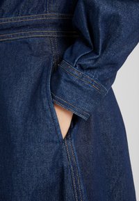 Dunkelblaue Jeansjacke mit einer Tasche, hochgerollten Ärmeln und sichtbaren orangefarbenen Nähten. Glatte Textur und lässiges Design.