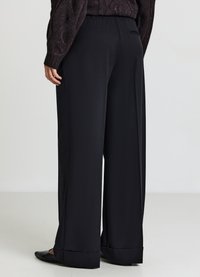 Schwarze Hosen mit weitem Bein, glatter Textur, elastischem Bund und umgeschlagenem Saum. Detail der Gesäßtasche auf der linken Seite sichtbar.