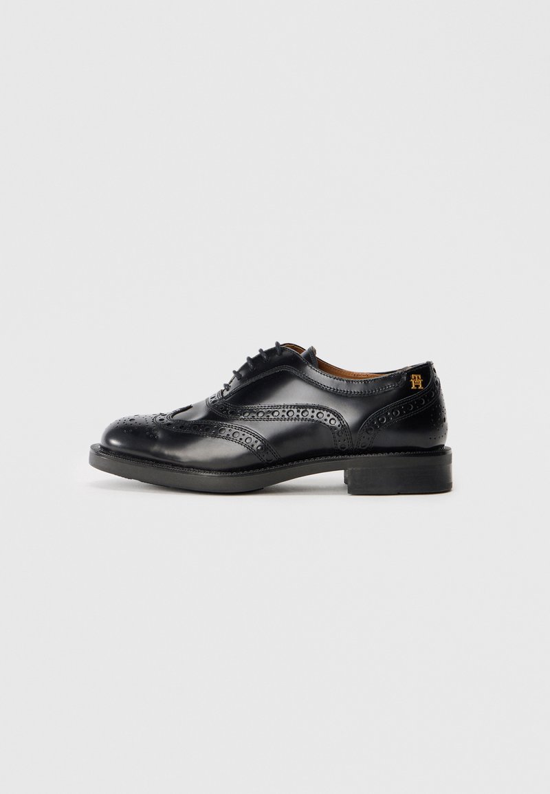 Black leather oxford shoe featuring perforated accents, a rounded toe, and a stacked heel. Smooth texture with contrasting brown inner lining.