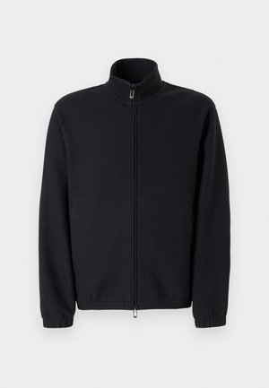 Melnā džemperis ar rāvējslēdzēju, izgatavots no strukturēta auduma, ar augstu apkakli un elastīgiem aprocēm, taisna griezuma un bez redzamām rotājumiem.