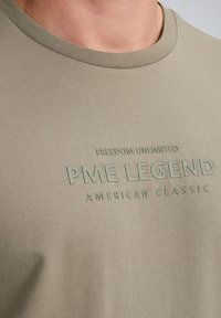 Olivově zelené bavlněné tričko s vyvýšeným textem: "NEOMEZENÁ SVOBODA PME LEGEND AMERICAN CLASSIC", s kulatým výstřihem a hladkým povrchem.