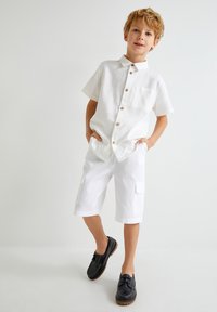 Camisa de lino blanca de manga corta con botones de madera y un bolsillo en el pecho, combinada con pantalones cortos cargo blancos con bolsillos. Zapatos negros.