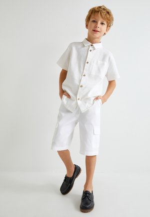 Camisa de lino blanca de manga corta con botones de madera y un bolsillo en el pecho, combinada con pantalones cortos cargo blancos con bolsillos. Zapatos negros.