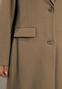 Primo piano di un cappotto marrone con due bottoni e una tasca a patta, che mostra parte della mano di una persona di lato.