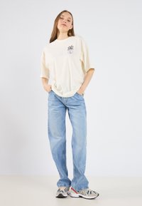 Camiseta crema con gráfico de palmera, combinada con jeans rectos de color azul claro y zapatillas multicolores, de pie sobre una superficie neutra.